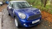 A blue MINI Cooper with a white roof is parked on a wet, leaf-strewn road beside a curb. The car's license plate reads "MD14 ULO." It is surrounded by autumn trees with partially bare branches and scattered fallen leaves. Other vehicles are visible in the background.