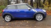 A blue two-door MINI hatchback with silver trim and a white roof is parked on a leaf-strewn driveway. The vehicle, adorned with silver alloy wheels, is positioned side-on. In the background, there is a wooded area with bare trees and scattered autumn leaves.