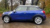 A blue MINI two-door compact car with a white roof and black accents is parked on a wet, leaf-strewn pavement. The car features silver alloy wheels and is positioned side-on, facing left. Bare trees with fall foliage are in the background, under an overcast sky.