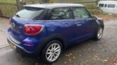 A metallic blue MINI Cooper with a black roof is parked on a wet road covered with fallen autumn leaves. The car has distinctive rounded taillights and silver alloy wheels. In the background, there is a wooden fence, greenery, a house with a moss-covered roof, and a white van.