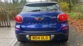 A blue MINI Paceman Cooper with the license plate "MD14 ULO" is parked on a wet concrete driveway. The surrounding area includes a wooden fence on the left, leaf-strewn ground, and trees with autumn foliage in the background. The car's rear lights are illuminated.