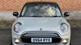 A front view of a silver Mini Cooper Diesel 5-Door with a UK number plate reading "OV64 RYX." The car features large circular headlights, fog lights, and a black roof. In the background, a brick wall and sparse vegetation are visible. The sky is beautifully reflected in the car's windshield.