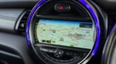 A car dashboard's circular display shows a GPS navigation map. The route is traced in pink, passing through locations like Stafford. The map is surrounded by a blue LED ring. Various control buttons are located below the screen, including volume and tuning knobs, all embedded into the diesel-fueled 2014 Mini Cooper's dashboard.