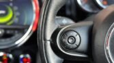 Close-up view of a Mini Cooper's steering wheel, focusing on the control buttons on the right side. The buttons include options like "LIM" (limit) and "RES" (resume). In the background, parts of the dashboard with a speedometer and other controls are visible but out of focus.