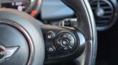 Close-up of a car steering wheel featuring multiple control buttons on the left side. Buttons include a phone icon, volume controls, and directional arrows with an "OK" button in the center. The dashboard and speedometer with a hint of orange lighting are partially visible in the background of this 5-Door Diesel Mini Cooper.