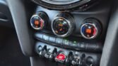 Close-up of a Mini Cooper's climate control panel. Three circular dials display temperature settings of 28 degrees, with the middle dial set to 'Auto'. Below the dials are toggle switches for max A/C, heated seats, and a red engine start/stop button. The 5-Door diesel model features a sleek black and shiny panel.