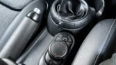 A close-up view of a 2014 Mini Cooper’s center console features a round gear selector with a leather cover, a circular control knob with buttons labeled "Map," "Nav," "Options," and "Back," and an adjacent handbrake lever. This 5-door diesel model showcases a primarily black console with grey accents.