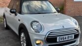 A 2014 Mini Cooper D with a black roof is parked on a driveway. The license plate reads "OV64 RYX." The car's right headlight is on, and there is a "NO PARKING" sign on the brick wall in the background. The sky's reflection is visible on the car's windshield.