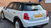 A white Mini Cooper 5-Door hatchback with a black roof is parked on an asphalt surface near a brick wall. The vehicle's rear end, including its license plate "OV64 RYX," is visible. The car has red taillights and a single exhaust pipe. There is a sign on the wall that reads, "NO PARKING.