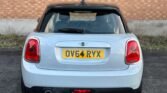 Rear view of a white MINI Cooper 5-Door Diesel with the license plate "OV64 RYX" displayed prominently. The car is parked on a paved surface in front of a brick wall. The left taillight is illuminated, and a small amount of exhaust smoke is visible near the tailpipe on the left side.