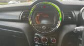 Close-up of the MINI car dashboard showing a central infotainment system. The display screen is tuned to a radio station called "D1 National Capital DANCE." Surrounding the display is a circular LED light ring, and below it are controls for air conditioning and other settings.