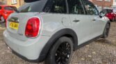 A light gray MINI Cooper with a black roof and black rims is parked on a gravel driveway in front of suburban houses. The license plate reads "Y165 GNV." The vehicle's rear and left side are visible, showcasing red tail lights and rain droplets on the car's surface.