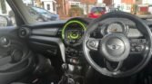 Image of a MINI's interior taken from the driver's perspective. Features include a black steering wheel with controls, a central infotainment screen with green backlighting, and various dials and controls on the dashboard. Outside, a residential area with brick houses and parked cars is visible.