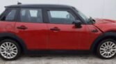 A red MINI Cooper with a black roof is shown in a side view. The vehicle, boasting four doors, is parked on a concrete surface in front of a plain white wall. The front right fender and bumper appear to be damaged. Its silver wheels gleam, and the windows are tinted for added privacy.