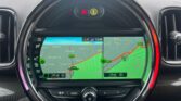 A MINI car navigation system displays a split-screen map view with traffic information. The left side shows a zoomed-out map and the right side a zoomed-in section. Alerts and waypoints are marked with icons. Below the screen, physical buttons for controlling the interface are visible. The time reads 12:46.