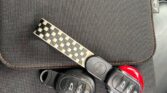 A black leather car seat serves as the backdrop for a set of MINI car keys placed on a black fabric pouch with brown stitching. The keys feature a checkered keychain and multiple buttons. The fabric pouch displays a small white logo in the shape of a stylized wing near the upper right corner.
