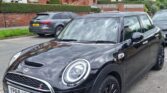 A black MINI Cooper S with a license plate reading "YP68 UTK" is parked on a residential street. The car features a prominent grille, large circular headlights, and black alloy wheels. Brick houses with red and white facades line the street, and a gray car is parked behind the MINI.