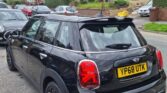 A black MINI Cooper with the license plate YP68 UTK is parked on the side of a residential street. The car is oriented rear three-quarters to the viewer, showcasing its taillights and dual exhaust. In the background, there are other parked cars, brick houses, and a cloudy sky.