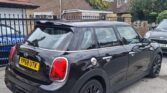 A black, four-door MINI Cooper hatchback with a yellow license plate reading "YP68 UTK" is parked on a residential street in front of a brick house with a tiled roof and leafy surroundings. Other cars are visible in the background. The overall scene is suburban and peaceful.