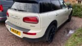 A white MINI Cooper Clubman parked on a gravel driveway. The car has a black roof and black-accented windows and wheels. The tailgate reads "Clubman" and "MINI Cooper" and features distinctive rectangular taillights. The driveway is next to a brick building and green shrubbery, with a grey van parked adjacent to the left.