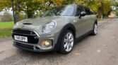 A gray MINI Clubman Cooper S with a license plate reading "FA19 OPP" is parked on a path surrounded by autumn foliage. The car features a black roof and side mirrors, sporty front grille, and large silver alloy wheels. The background shows trees with yellow and green leaves and a grassy area.