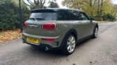 A grey MINI Clubman is parked on a tree-lined street with yellow and green leaves on the ground. The car's rear, showcasing a FA19 OPP license plate, dual exhausts, and distinct rear lights, is in focus. Nearby, a lamp post and hedges are visible on an overcast day.