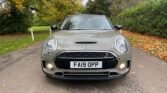 A grey MINI Clubman with a black roof is parked on a leaf-strewn driveway, surrounded by autumn trees and greenery. The car's headlights are on, and its license plate reads "FA19 OPP." The scene captures the essence of fall with leaves scattered on the ground.