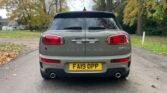 A gray 2019 MINI Clubman Cooper S is parked on a paved driveway surrounded by autumn foliage. The rear view of this estate car shows its distinctive split rear doors, red taillights, dual exhaust pipes, and a yellow license plate with the registration number FA19 OPP. Trees and a house are visible in the background.