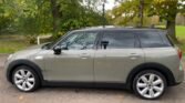 A light olive-green 2019 MINI Clubman Cooper S is parked on a road with its side facing the camera. The car has tinted windows, silver door handles, and multi-spoke alloy wheels. In the background, there are trees with autumn foliage, a green grassy area, and a black lamp post.