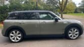 A gray MINI Clubman is parked on an asphalt road with a backdrop of autumn trees and greenery in a suburban area. The car's side profile showcases its five-door design with silver rims. A streetlamp and fallen leaves are visible, indicating a calm, fall day for this stylish Petrol Estate.