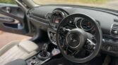 Interior of a 2019 MINI Clubman featuring a black leather steering wheel with the MINI logo at the center. The dashboard includes a large central display, round air vents, and various control buttons. Beige leather seats and lush green scenery are visible through the windows of this Petrol Estate.