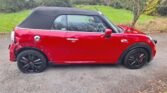 A red 2020 Mini John Cooper Works convertible with a black soft top is parked on the side of a road. The car appears damaged on the rear wheel arch, with visible dents and missing paint. Set against a lush green field and trees, this repairable vehicle features sleek black alloy wheels.