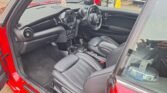 The interior of a red Mini John Cooper Works Convertible is shown with the driver's side door open. The car features black leather seats, a multifunction steering wheel, and a dashboard with circular air vents. Several controls and cup holders are visible between the driver's and passenger's seats.