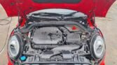 The image depicts the open hood of a red Mini John Cooper Works, revealing its engine compartment. The black engine cover prominently displays the Mini logo. Visible components include the air intake, battery, and various hoses and wires. The car is parked outdoors on a wet surface.