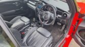 A view of a 2020 Mini John Cooper Works Convertible's interior from the driver's side reveals black leather seats. The steering wheel, laden with multiple controls, complements a dashboard filled with instruments and a central display. The exterior boasts bright red paint, with a small air freshener hanging from the rearview mirror.