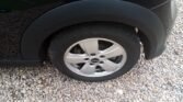 Close-up of a MINI's rear wheel on a gravel surface. The car boasts a black exterior with visible tire tread and sleek, silver alloy wheels. The surrounding background includes small, uniformly sized pebbles.