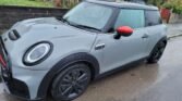 A grey 2022 MINI COOPER S with a black roof and red side mirror caps is parked on a wet, residential street. The car has black wheels and rain droplets on its surface, suggesting recent rainfall. Despite its 15000 miles, it appears undamaged. Residential houses and a stone wall are visible in the background.