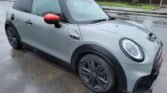 A sleek, gray MINI Cooper S with black alloy wheels and red side mirrors is parked on a wet street. The car's modern design features rounded headlights and a distinct front grille, despite some minor damage. Residential houses with brick walls and fences are visible in the background on a cloudy day.