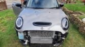 A gray 2022 MINI COOPER S with its front bumper removed, revealing the radiator and internal components. The damaged car, which has clocked 15,000 miles, is parked on a grassy area in a residential neighborhood with wooden fences and houses in the background. One of the car's doors is open.