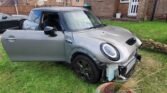 A gray MINI Cooper S with significant front-end damage is parked on a grassy lawn, partially resting against a potted plant. The front bumper is detached, and the driver's side door is open. A brick house with a white-framed door and windows is visible in the background, highlighting the unfortunate scene.