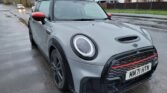 A grey 2022 MINI COOPER S with black and red accents is parked on a wet road. Its license plate reads "MM71 HTN." Nearby houses and a car driving in the opposite direction are visible. The area appears to be a suburban neighborhood with overcast weather and wet pavement from recent rain, showing signs of light damage at 15000 miles.