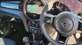 Interior of a MINI COOPER S featuring a steering wheel with the Mini emblem, a digital dashboard displaying speed and other metrics, and a central touchscreen showing a map. Below the screen are various control buttons and knobs. The car has 15000 miles on it, with a manual transmission gear stick and cup holder in view.