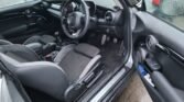 The image shows the interior of a modern car from the driver's side with the door open. The seats and dashboard are upholstered in black material. The sporty 2022 MINI COOPER S boasts chrome accents on the steering wheel, pedals, and dashboard. It appears to be wet outside, indicating recent rain.