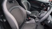 Interior view of a MINI COOPER S featuring two front seats with black upholstery and gray accents. The dashboard and steering wheel are visible, with red trim details on the door panel. The car windows have rain droplets, indicating wet weather outside. This compact car has headrests and armrests, with 15,000 miles on the odometer.