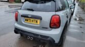 A grey MINI COOPER S with 15,000 miles on the clock is parked on a wet street, facing away from the camera. The car has a visible UK registration plate reading "MM71 HTN." Its design features include a rear spoiler, distinctive tail lights, and dual exhaust pipes. The surrounding area appears damp, likely due to recent rain.