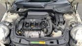 A photo of a MINI's engine bay with the hood open, revealing the engine and various components. The engine cover displays the manufacturer's logo. Visible parts include the air intake, battery, fluid reservoirs, hoses, and belts. The surrounding area is slightly dusty, indicating regular use.