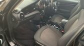 Interior of a black MINI car showing the front seats, dashboard, and steering wheel. The seats are black with grey fabric center panels. The dashboard features a circular center console with various controls and a display screen. The steering wheel has multiple buttons and controls.