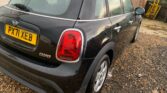 A black MINI Cooper with a white roof is parked on a gravel driveway. The vehicle has a UK number plate reading "PX71 XEB". The car is wet, suggesting it may have recently rained. In the background, there is a wooden shed, a yellow hand truck, and a fence.