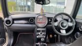         Interior of a modern MINI Cooper car showing black and grey front seats, a compact dashboard with circular air vents, a large central speedometer, and a multifunctional steering wheel. The gear shift is visible, and there is a small rearview mirror hanging from the ceiling.