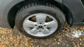 Close-up of a silver alloy wheel on a black MINI, surrounded by damp, brown, and tan gravel. The tire is branded with "Hankook" text and appears wet from rain. The wheel has five circularly arranged holes and four visible wheel nuts. Some small white foam patches can be seen near the tire.